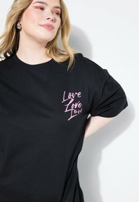 Czarna bawełniana koszulka z różowym nadrukiem graficznym z napisem "Love" w różnych orientacjach na lewej stronie klatki piersiowej. Krótkie rękawy.