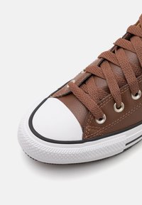 Hnědé kožené tenisky s bílým gumovým krytem špičky a podešví, s kulatými kovovými očky, hnědými tkaničkami a texturovanými detaily po stranách.