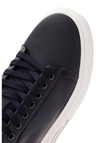Sneaker in pelle blu navy con punta arrotondata, suola in gomma bianca e lacci intrecciati attraverso occhielli in argento. Texture liscia con dettagli minimi.