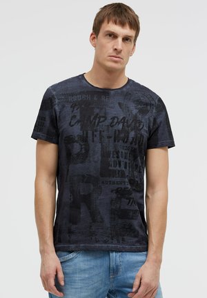 Young man with short brown hair wearing a dark graphic T-shirt and blue jeans standing against a plain light background.