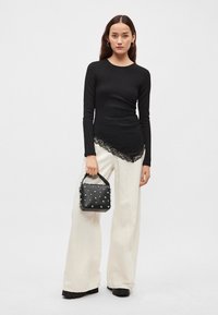 Schwarzes Langarmoberteil mit Spitzenbesatz, kombiniert mit weitem weißen Hosen. Hält eine kleine schwarze Tasche mit Nieten, trägt Creolenohrringe.