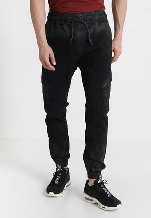 Schwarze Jogginghosen mit elastischem Bund und Kordelzug. Mit Seitentaschen und schmal zulaufenden Beinabschlüssen. Getragen mit schwarzen Sneakern und einem bordeauxfarbenen Oberteil.
