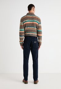 Suéter de punto marrón con rayas y acentos en azul, naranja y crema; pantalones azul marino; persona vista desde atrás. Textura plana y suave.