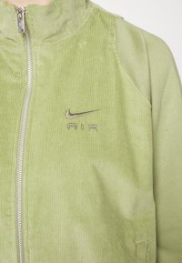 Lys grøn kordfløjlsjakke med høj krave, sølv lynlås og broderet Nike-logo på brystet. Glat tekstur med lodret ribbede detaljer.