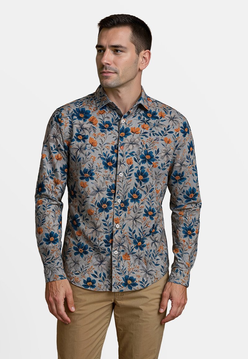 Man in een grijs overhemd met knopen, versierd met een blauw en oranje bloemenpatroon, en een beige broek, staande en naar de zijkant kijkend.