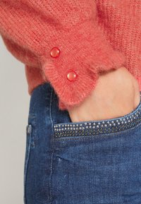 Koraal kleurige fuzzy breisweater met twee rode knopen bij de manchetten, gecombineerd met blauwe denim jeans met een bejeweelde zakrand.