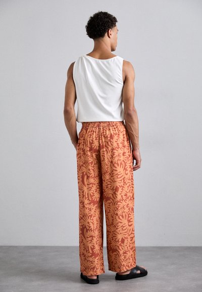 Pantalons amples orange avec un motif floral jaune, ceinture élastique et sandales noires, associés à un débardeur blanc.
