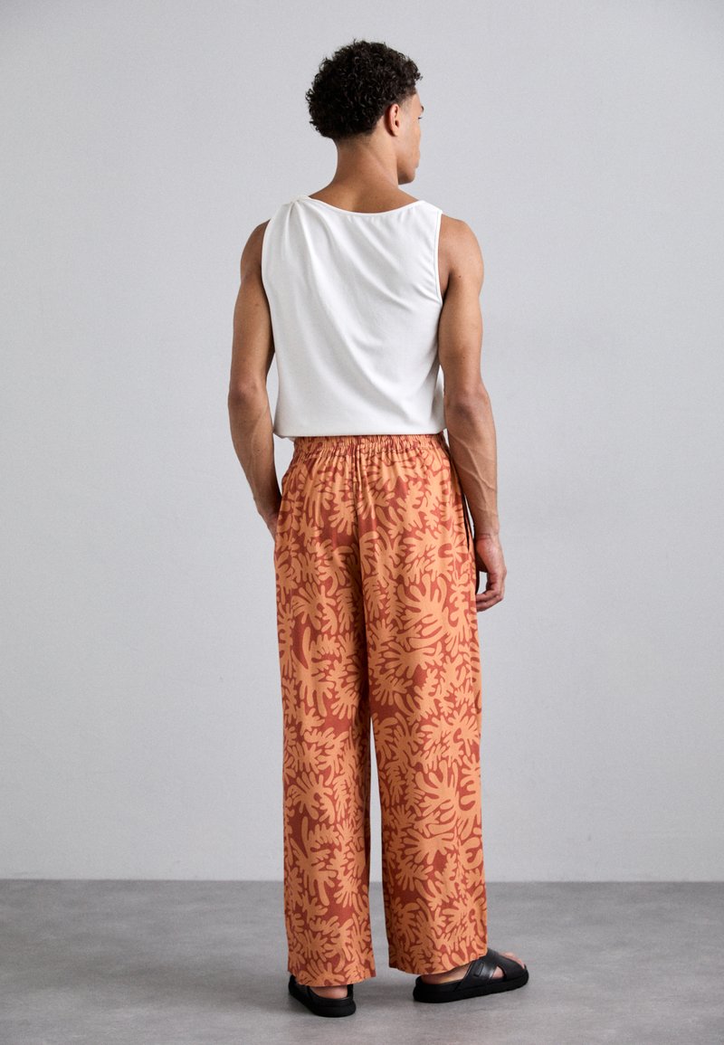 Loose-fitting orange pants with a yellow floral pattern, elastic waistband, and black sandals, paired with a white tank top.