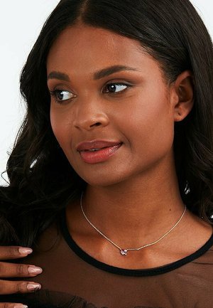 Silberne Kettenhalskette mit einem kleinen rosa Edelsteinanhänger, getragen von einem Modell mit dunklem Haar, vor einem schwarzen transparenten Oberteil.