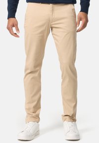 Beige bomuldsbukser med slim fit, der har lynlåsgylp og sidelommer, parret med hvide sneakers. Stoffet er glat og let.