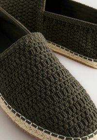 Mørkegrønne hæklede slip-on sko med en tekstureret overflade, der har en sål i naturfiber og kontraststikninger langs kanten.