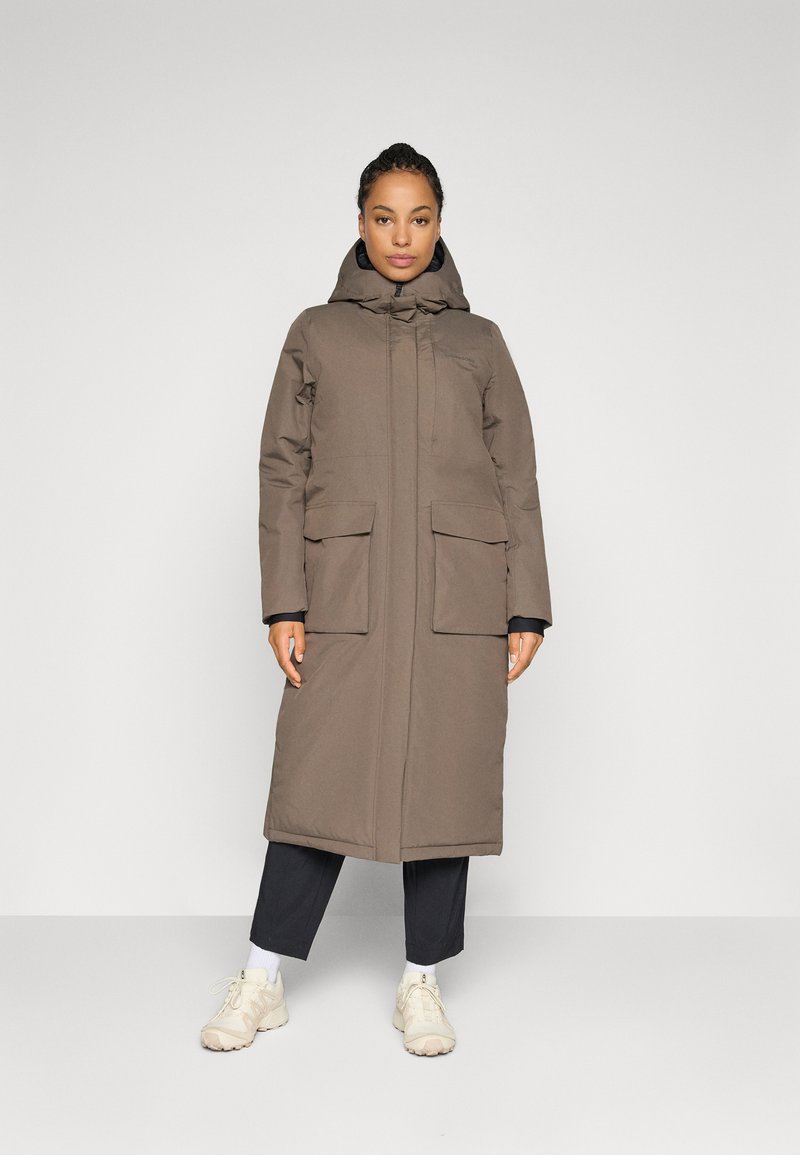 Donna che indossa un lungo cappotto invernale marrone con cappuccio e grandi tasche frontali, pantaloni neri, calzini bianchi e sneaker beige, in piedi su uno sfondo grigio.