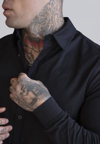 Fekete gombos ing, sima textúrával, gallérral és hímzett logóval a mellkason. Lekerekített mandzsettákkal rendelkezik és látható tetoválások vannak a kezén.