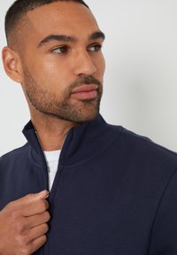 Mann mit gestutztem Bart, der eine navyfarbene Zipfeljacke trägt, den Reißverschluss haltend, zur Seite schauend vor einem schlichten Hintergrund.