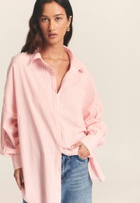 Pink striped oversized button-up shirt with a collar, long sleeves, and a chest pocket, paired with blue denim jeans.
