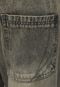 Κοντινό πλάνο μιας αποχρωματισμένης πίσω τσέπης από μαύρο τζιν με εμφανείς ραφές και μεταλλικά καρφιά στις πάνω γωνίες.