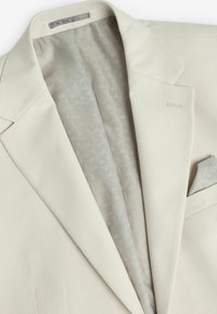 Blazer beige clair avec un tissage texturé. L'intérieur présente un motif géométrique en gris clair. Comprend un mouchoir de poche de couleur assortie.