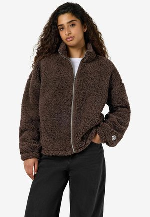 Femme aux longs cheveux bouclés portant une veste polaire marron zippée et un pantalon noir, debout avec une main dans la poche sur fond blanc.