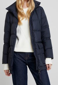 Chaqueta acolchada azul marino con cierre frontal, gran cuello y bolsillos laterales. Presenta un diseño acolchado y puños acanalados, llevada sobre un suéter color crema.