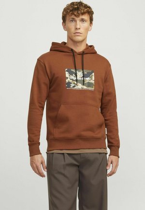 Brun hoodie lavet af bomuld med en frontlomme, snor i hætten og et grafisk print. Parret med grå bukser. Enkel design.