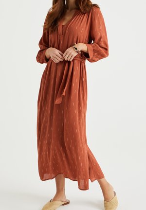 Kvinde iført en lang rustfarvet kjole med subtile mønstre, bælte i taljen og beige vævede slip-on sko.