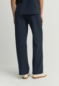 Pantaloni largi de culoare albastru marin, confecționați dintr-un material moale, având un design simplu și o talie elastică, asortați cu pantofi de o culoare deschisă.