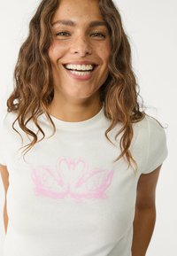 Biała koszulka z krótkim rękawem z różowym motywem łabędzia na klatce piersiowej, wykonana z miękkiego materiału, z klasycznym okrągłym dekoltem.