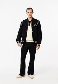 Chaqueta varsity negra con rayas blancas, parche de logo verde. Polo blanco con ribetes debajo, pantalones negros y zapatos beiges. Sosteniendo una bolsa pequeña.