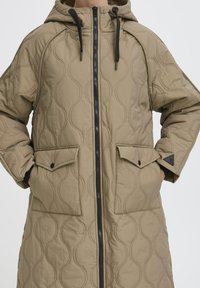 Gepolsterte beige Parka mit Kapuze, ausgestattet mit einem schwarzen Reißverschluss, zwei vorderen Taschen und gerippten Bündchen, die ein Wellenmuster auf dem Stoff zeigt.