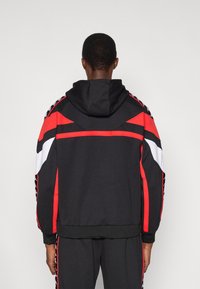 Črna kapucar s kapuco s rdečimi in belimi poudarki, ki ima na ramenih drzne geometrijske vzorce in rebrasti rob dna.