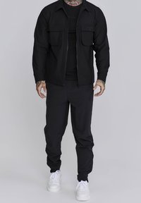 Svart zip-up jacka med två bröstfickor, i kombination med svarta smala byxor och vita sneakers. Tyget verkar slätt med en åtsittande design.