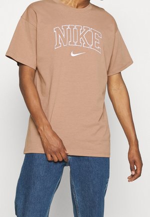 Persoon met een bruine Nike T-shirt met wit logo en blauwe spijkerbroek, staand tegen een effen lichte achtergrond.