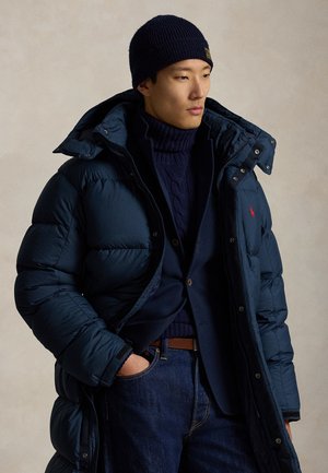 Mann trägt eine marineblaue Mütze, einen marineblauen Rollkragenpullover, einen marineblauen Blazer und eine marineblaue Steppjacke mit Jeans, steht vor einem neutralen Hintergrund.