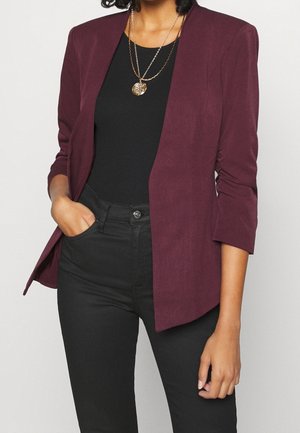 Vrouw draagt een bordeauxrode blazer over een zwart topje en zwarte broek, met twee gouden kettingen en één hand in haar zak.