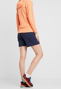 Sudadera con capucha naranja, pantalones cortos azul marino y zapatillas deportivas multicolores. La sudadera tiene una tela texturizada y un ajuste relajado.
