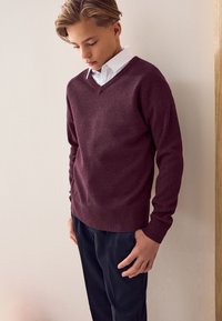 Burgundi színű V-nyakú pulóver puha anyagból, bordázott mandzsettákkal és szegéllyel, sötét nadrággal párosítva, semleges háttér előtt bemutatva.