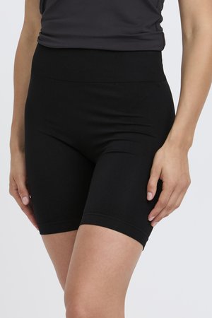 Femme portant un short de cycliste noir moulant taille haute et un haut noir, debout les bras détendus le long du corps devant un fond uni.