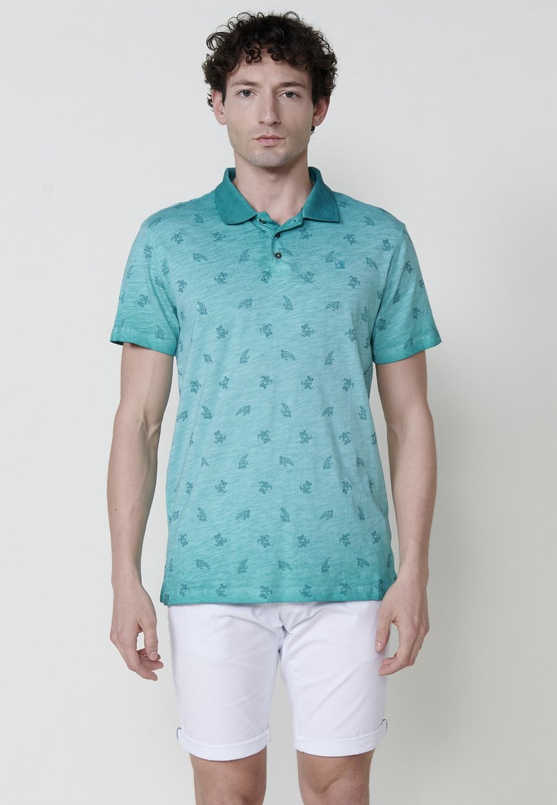 Rövidujjú poloing türkiz színben, apró sötétzöld mintázattal, fehér felhajtott rövidnadrággal párosítva.