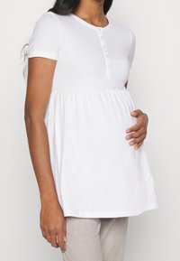 Hvid kortærmet skjorte med knaplukning ved henley-halsen og indsnævret talje, lavet af blødt bomuldsstof, designet til komfort under graviditet.