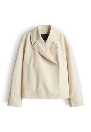 Veste courte beige avec larges revers, manches longues et poches verticales à coutures sur le devant, présentée sur fond blanc uni.