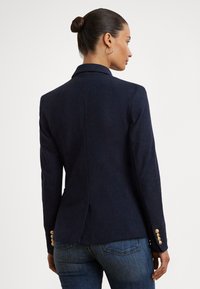 Blazer azul-marinho com corte ajustado, lapelas estruturadas e detalhes de botões dourados nas mangas. Tecido texturizado, folga nas costas.