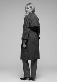 Trench-coat noir à taille ceinturée, avec un large col et des manches retroussées, associé à un jean droit et des escarpins à bouts pointus.