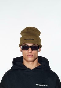 Olíva zöld kötött sapka hajtott mandzsettával és logóval; fekete kapucnis hoodie elülső logóval; elegáns fekete napszemüveg; semleges háttér.