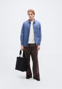 Τζιν πουκάμισο με τσέπη στο στήθος, λευκό T-shirt από κάτω, καφέ παντελόνι με φαρδύ πόδι και μαύρη τσάντα ώμου με κοντά χερούλια.