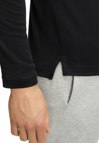 Schwarzes langärmliges Oberteil mit Rundhalsausschnitt und Seitenschlitzen, aus glattem Stoff und mit genähten Bündchen, kombiniert mit grauen Sweatpants.