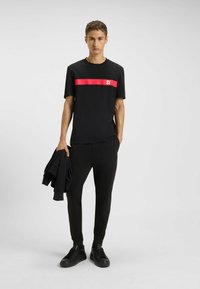 T-shirt nera con striscia orizzontale rossa e logo, abbinata a pantaloni jogger neri con accenti rossi. Il modello tiene in mano una giacca nera.