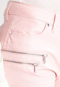 Los pantalones de mezclilla rosa cuentan con dos bolsillos decorativos con cremallera en la parte trasera, con una textura suave y acentos en hardware plateado en contraste.