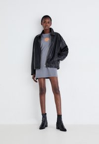 Black leather oversized jacket, gray dress with orange sun graphic. Model wears black heeled ankle boots, standing against a plain wall.