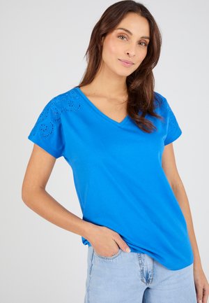 Vrouw met lang bruin haar, draagt een felblauw T-shirt met V-hals met kanten details op de mouwen en lichtblauwe jeans, hand in de zak.