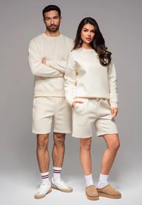 Des sweat-shirts et des shorts de couleur crème fabriqués à partir d'un tissu doux. L'homme porte un sweat-shirt à manches longues, tandis que la femme a un col rond et des poches.